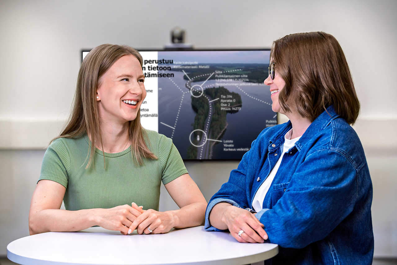 Kaksi henkilöä keskustelee ja hymyilee toisilleen. Taustalla olevalla näytöllä on esitys.