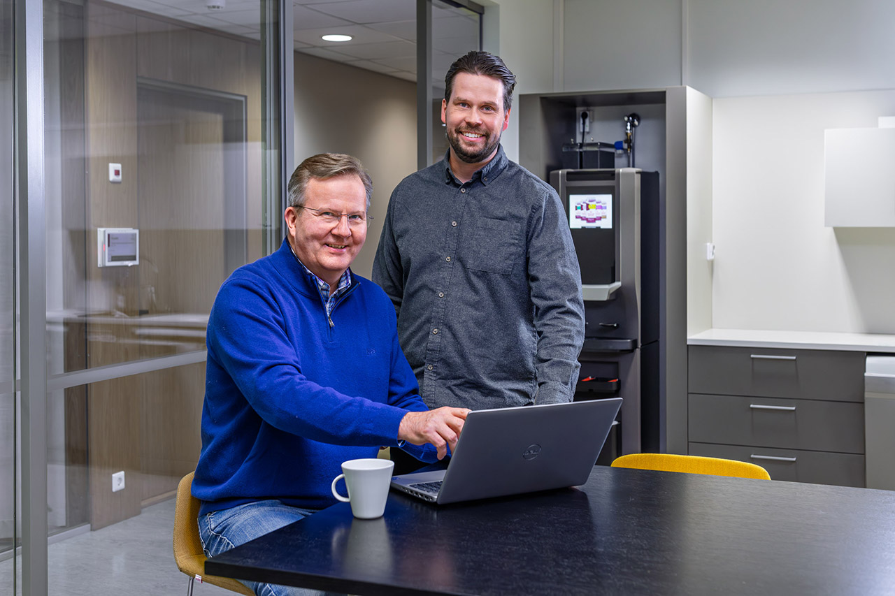 Kaksi henkilöä on pöydän ääressä, toisella on edessään tietokone. He katsovat kameraan.