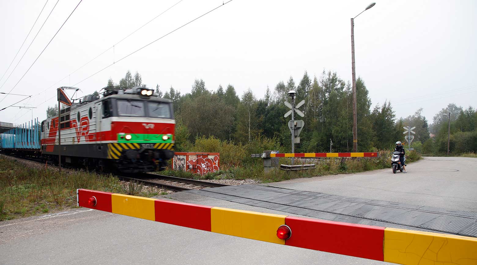 Tasoristeys, jossa puomit alhaalla, koska juna juuri tulossa. Toisella puolella mopoilija odottaa radanylitysvuoroaan.