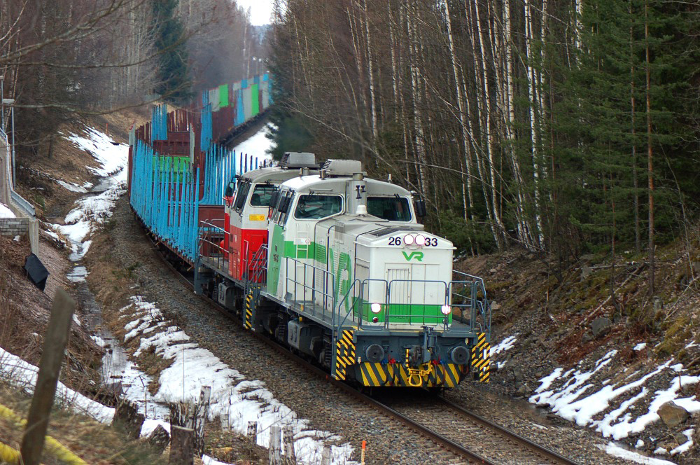 Kuvassa vihreävalkoinen veturi vetää  puunkuljetukseen tarkoitettuja tavaravaunuja keskellä metsää.