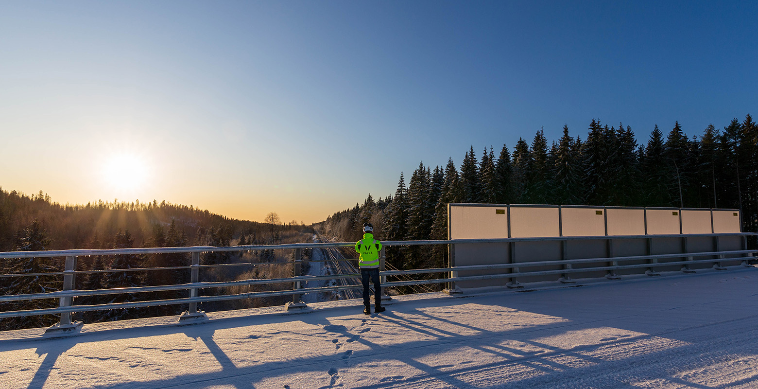 Letke-projektin projektipäällikkö seisoo uudella lumisella sillalla ja katselee alla olevaa rataa auringon laskiessa taustalla