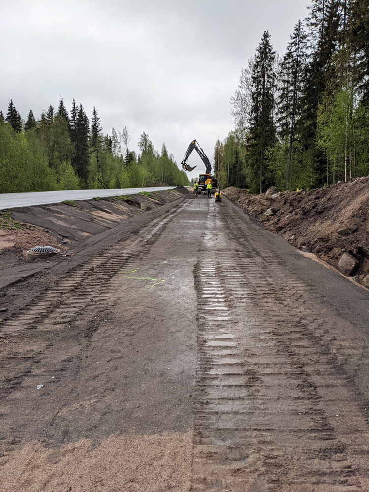 Kevyen liikenteen väylän pohjalle levitetty tasainen, kostea sorakerros, taustalla kaksi työntekijää ja kaivinkone.