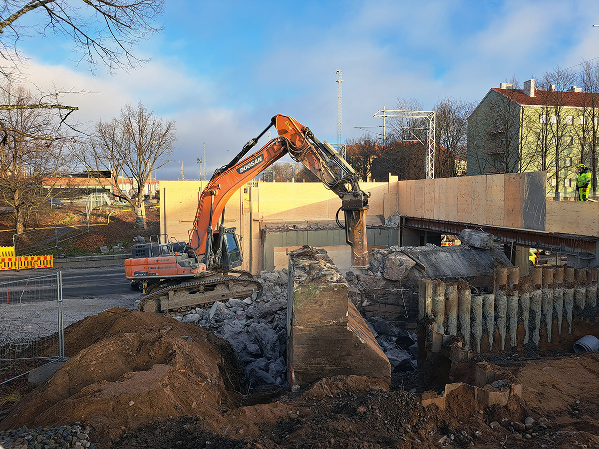 Kaivinkone purkaa vanhaa betonirakennetta työmaalla. Etualalla näkyy murskattua betonia ja maata, ja taustalla uutta siltaa. Oikealla työskentelee rakennustyöntekijöitä, ja taustalla näkyy asuinrakennuksia ja lehtipuita.