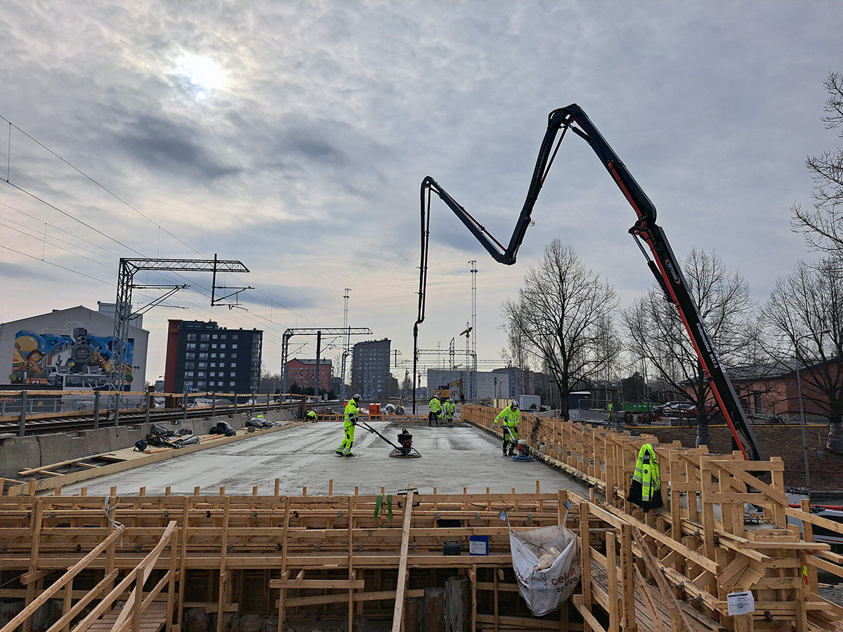 Työmaalla useat rakennustyöntekijät viimeistelevät juuri valettua betonipintaa sillan kannella. Betonipumppu ulottuu korkealle taivaalle, ja työntekijät käyttävät tasoituskoneita. Puurakenteinen valumuotti ympäröi siltaa, ja taustalla näkyy kerrostaloja, junarataa ja nostureita.