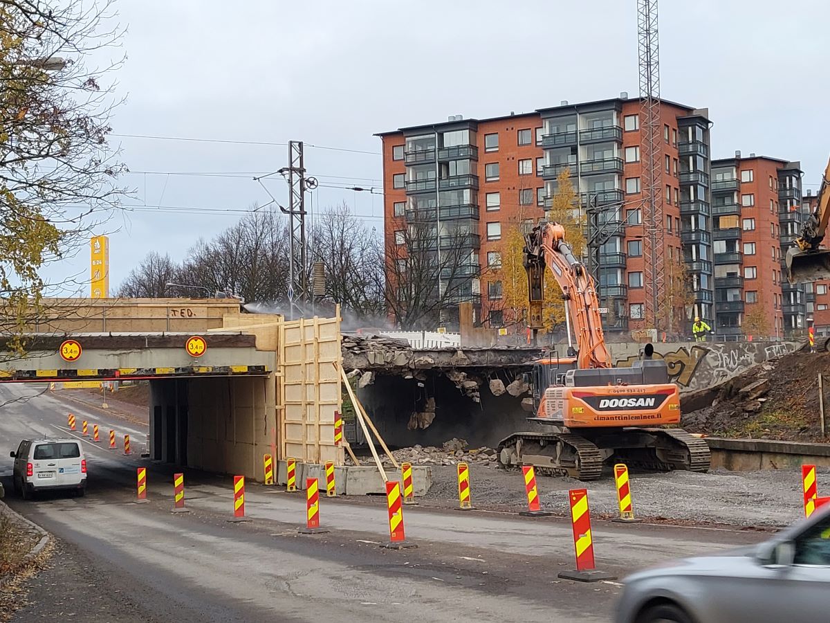 Työmaa, jossa kaivinkone purkaa vanhaa betonista rautatiesiltaa. Vieressä kulkee suojattu alikulku, jota autot käyttävät. Taustalla näkyy kerrostaloja ja syksyisiä puita.