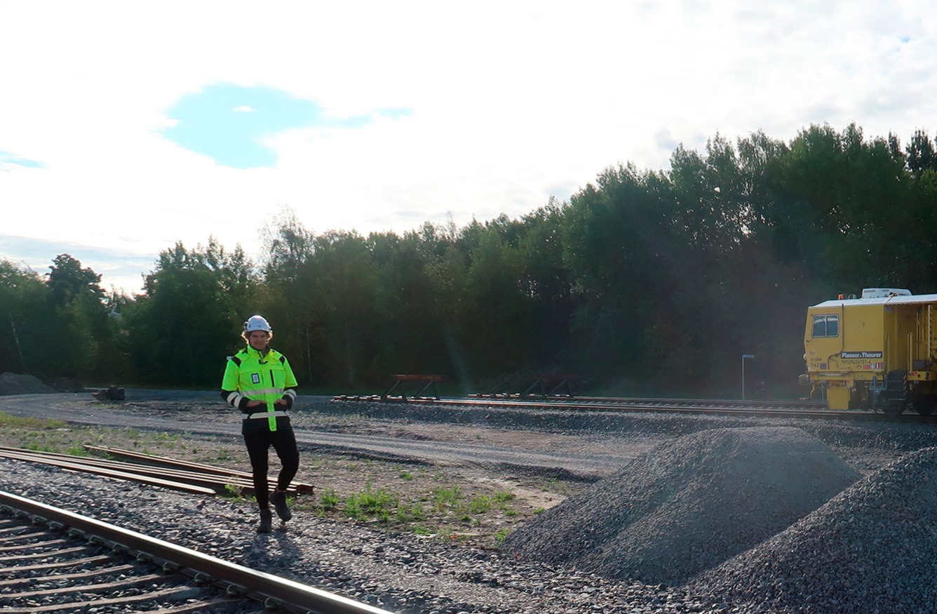 Rautatiealueella seisova henkilö huomioliivissä ja kypärässä, taustalla junan huoltokone ja sorakasoja.