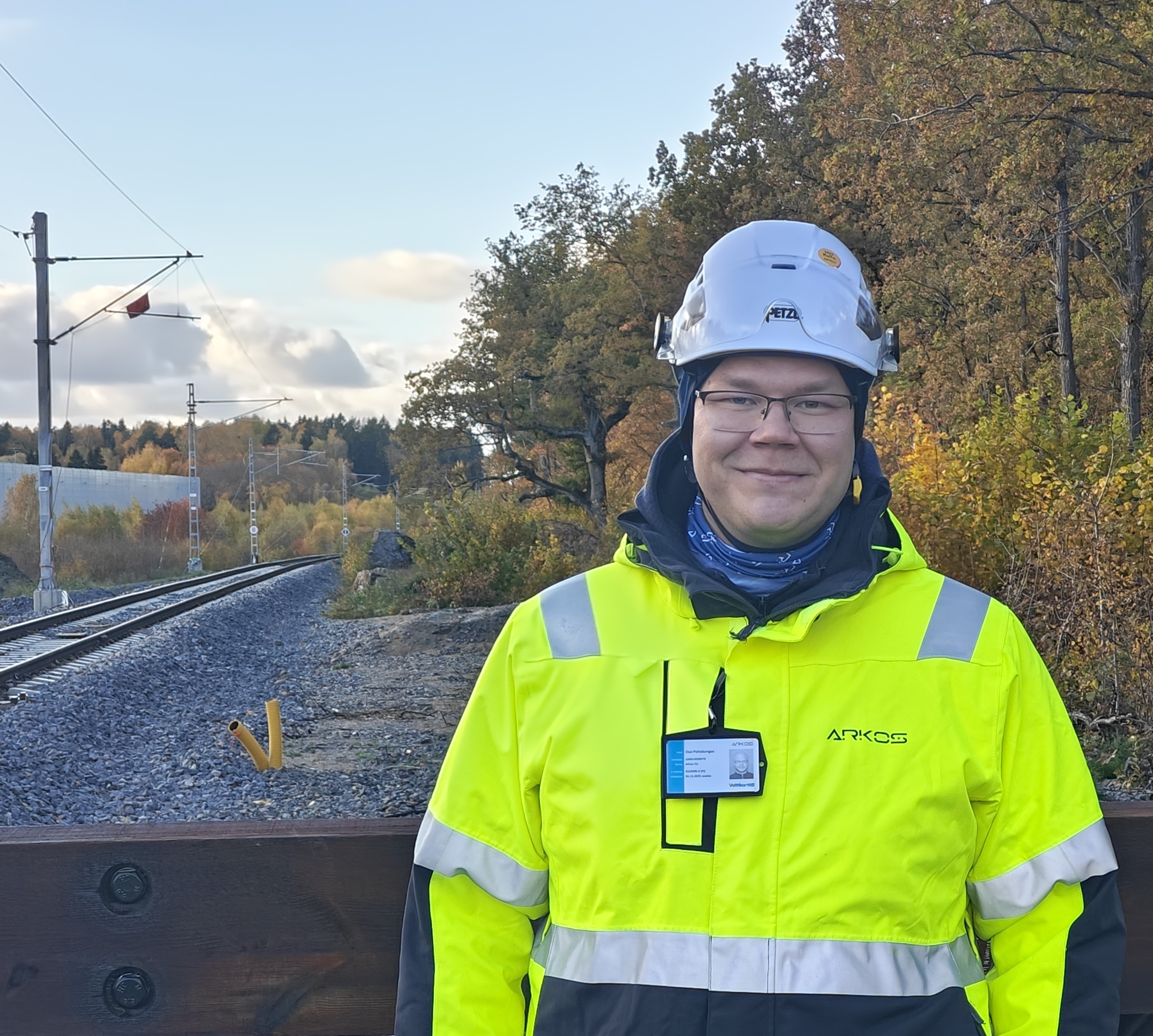 Mies rautatien vieressä syksyisessä maisemassa. Hänellä on päällään kirkkaan keltainen työvaate ja valkoinen suojakypärä. Taustalla näkyy rautatiekiskoja, puita, ja sininen taivas, jossa on pilviä.