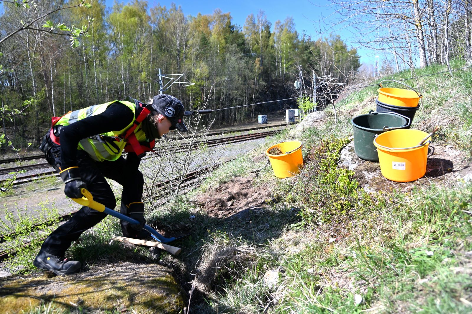 Työntekijä kaivaa maata lapion avulla rautatien vierellä. Hänellä on yllään turvavarusteet ja viereen on aseteltu kolme värikästä ämpäriä.