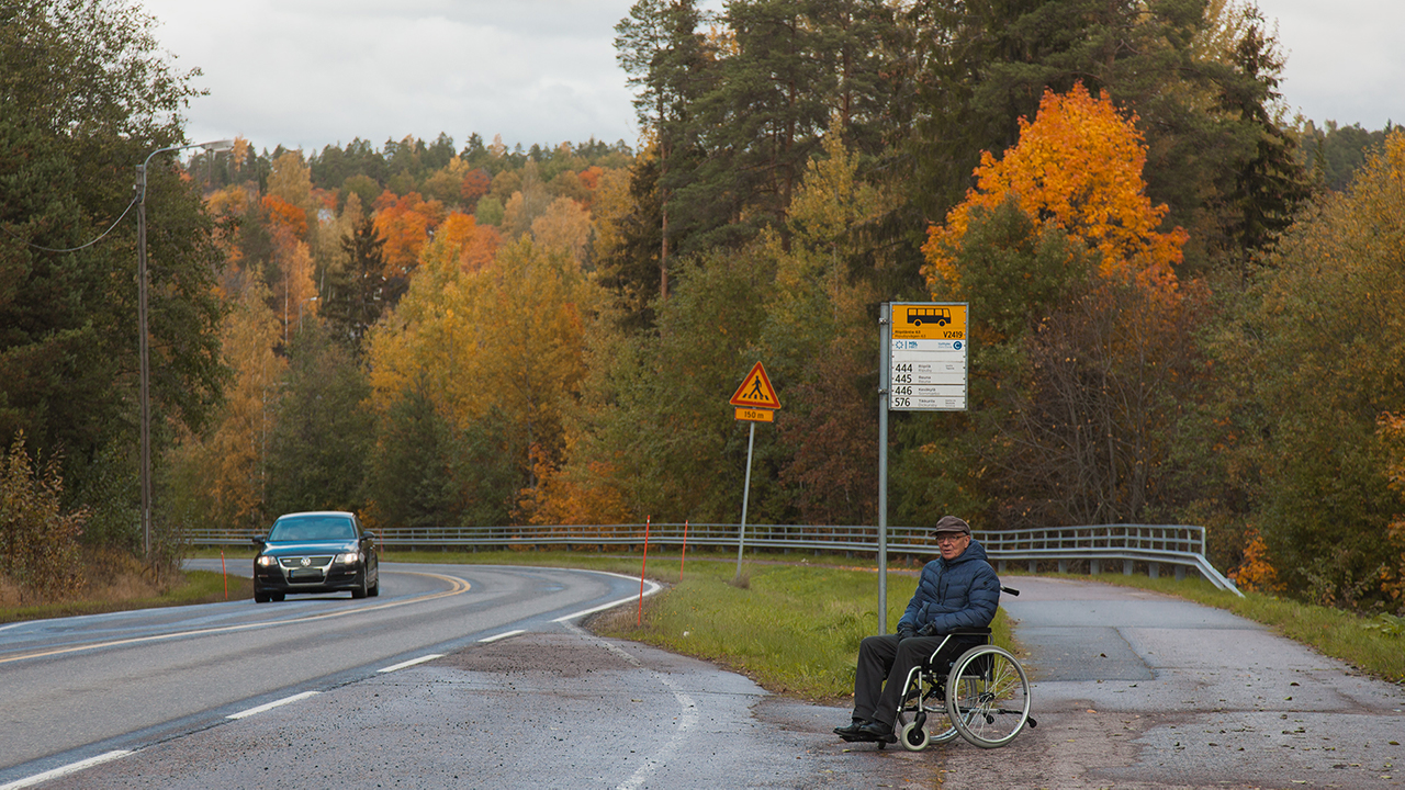 Iäkäs mies istuu pyörätuolissaan bussipysäkillä. Toisella puolella tietä musta auto ohittaa. Taustalla syksyinen metsä.